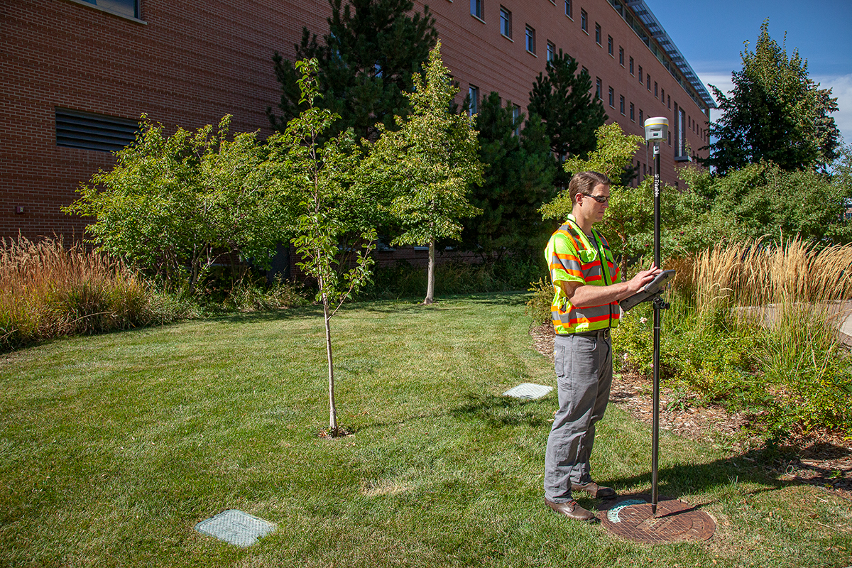 Trimble R12 Man standing surveying a manhole cover in the grass with a GNSS rover in a city setting