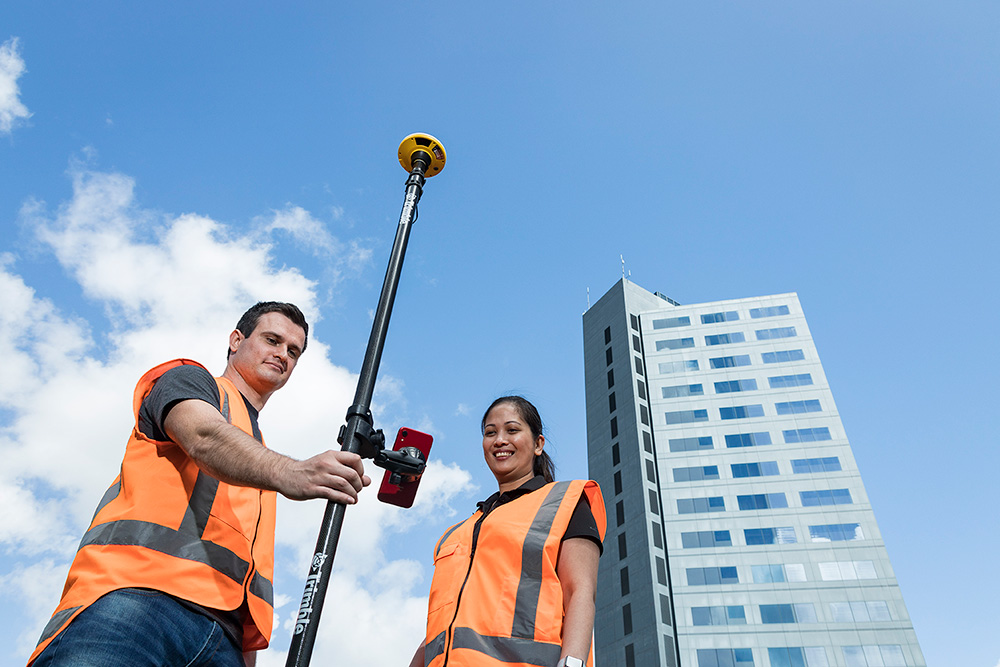 DA2:作为 Trimble Catalyst 定位服务核心的 GNSS 接收机 高层建筑旁一男一女真正查看安装在对中杆上的手机